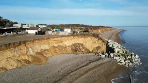 PA Media An aerial image looking inland from above the sea. A sandy cliff has been exposed by erosion, creating a bay. At either end, sea defences have been placed to try to protect the cliff. The rest of the village is visible behind, including a lake.