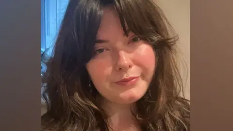 Chloe Humphries who has long dark wavy hair over her shoulders. Her head is tilted to her right and she has a serious expression on her face. 