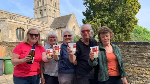 Ann Huckle Five women - one in a pink T-shirt - holding a bottle of fizz. The others are all holding a white plastic cup with a British flag on it, and are smiling for the camera. 
