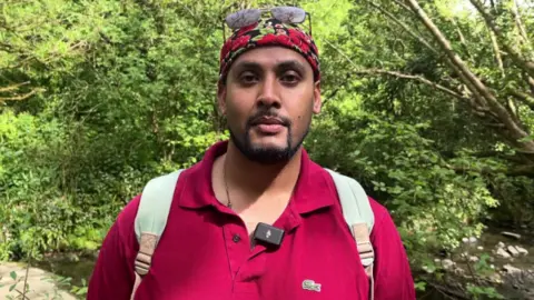 Hari is seen from his shoulders up. He is wearing a red T-shirt and a backpack. There is a woodland and river behind him.