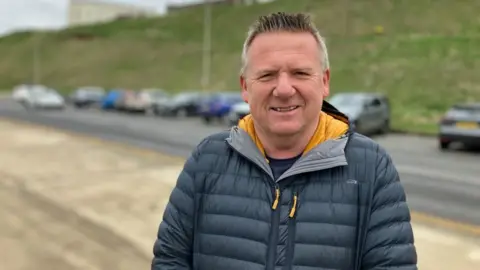 BBC/Seb Cheer A man, Fraser, smiles at the camera. He is wearing a warm coat. A line of parked cars is in the background.