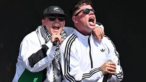 Two men wear white tracksuit tops. The man on the left has his hand over the shoulder of the man on the right. He holds a black microphone and wears a black and white scarf and a black baseball cap. The man on the right has on black sunglasses.