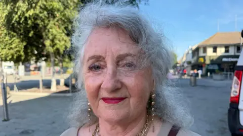Richard Knights/BBC Jenny Hopkins has shoulder-length grey hair. She is wearing golden jewellery and smiling in Clacton town centre.