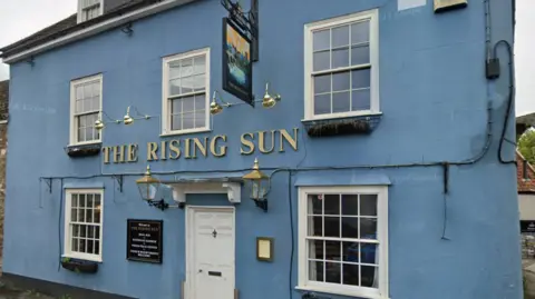 Google A picture of Mr Moore's pub called The Rising Sun. The name is in gold on a blue coloured building. The door and window frames are white.