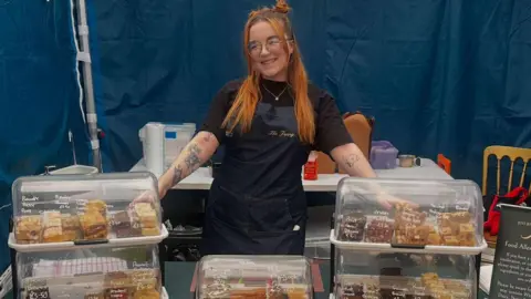 Just Jenny’s Jenny Merrick wears glasses and a dark apron. She has vibrant orange hair. She points to trays of cakes in clear boxes in a marquee with blue walls.