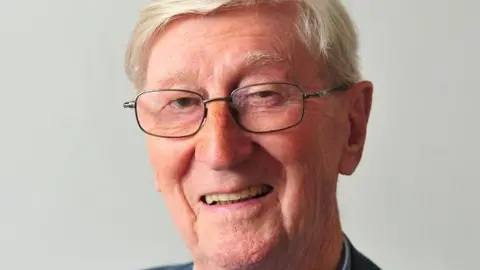The picture shows an older man sitting against a plain light background. He has short, neatly combed white hair and is wearing thin, rectangular glasses. He is smiling in a warm, relaxed way. 