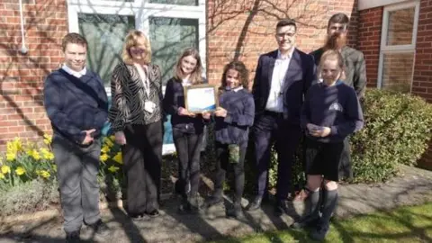 Headteacher Luke Dancer with Emma Wooldridge, SENCO, Councillor Bob Webb, and pupils from Copmanthorpe Primary School, standing in front of a red brick building on a path, with a flower bed of daffodils behind them and a grass lawn in front of them.