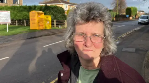John Devine/BBC Karen McFarling has grey hair in a bouffant style with gold framed glasses, she is wearing a purple fleece top, with a light green T-shirt underneath.
Behind her is a fire station with two large yellow charity collection bins for clothes and shoes, with no rubbish or any bags visible.