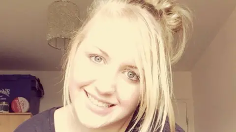 Family handout Abbi Smith sits in her bedroom smiling at the camera. She is in her 20's. She has light blonde hair pinned up in a loose bun and has very light blue grey eyes. Strands of blonde hair are falling down around her face. She has a black round neck top on and you can see items on top of her wardrobe behind her, along with a sparkly lampshade on the ceiling.