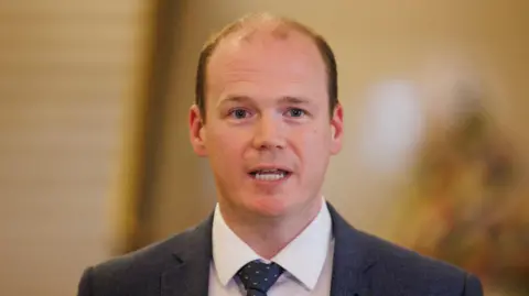 Gordon Lyons is standing in front of the camera and looks as though he is speaking. He is wearing a blue jacket with a spotted tie and a white shirt. He has fair brown hair which is thinning on the top