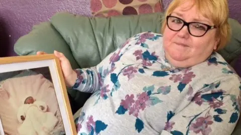 Llinos sitting on a couch holding a framed photo of a tiny baby in a white baby's gown and bassinette. Llinos is looking at the camera and has a sad look on her face. She has short blonde hair and wearing black rimmed glassed with a floral patterned top