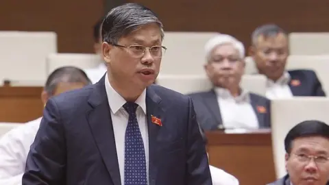 Supplied: Government of Vietnam A man in a suit, tie and glasses, with a small red badge pinned to his lapel, stands inside what appears to be a parliament building