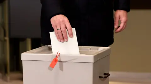 BBC A person in a black coat, hold a white piece of paper in their hand with a silver ring, and drops it into a grey box with a handle and red tag.