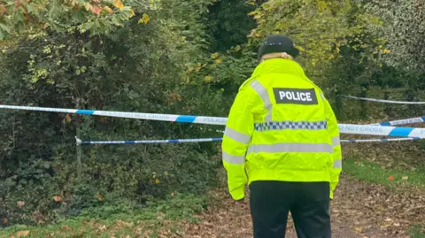 Jamie Niblock/BBC A police officer dressed in a high-visibility jacket and black trousers, standing in front of blue and white police tape. The tape cordons off a wooded area and a path with leaves on the ground.