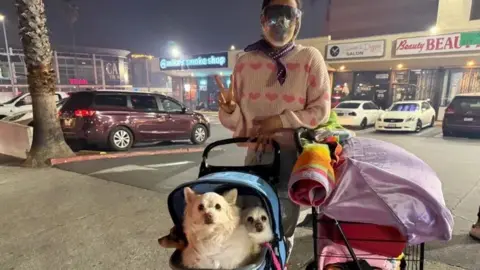 Crystal Hayes Anna Waldman makes a peace sign as she stands with her belongings and three dogs in a stroller. 