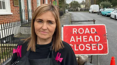 A woman with hair clips on her clothes standing in front of a red "Road Ahead Closed" sign.