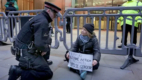 PA Media Greta Thunberg sitting on the pavement behind police barriers, holding a placard reading “I support Palestine Action prisoners. Oppose genocide,” as a police officer kneels to speak to her outside an office building in the City of London.