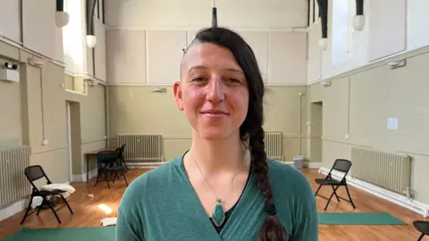 Ellen Knight/BBC Photograph of Burrows looking into the camera and smiling slightly. She has long, dark brown hair worn in a plait, and has the right side of her head shaved. She is wearing a dark green, long sleeved top and a necklace with a green leaf-shaped pendant. Burrows is pictured inside a hall with green and white painted walls with a wooden floor. Behind her are two black fold-away chairs and two dark green yoga mats. 