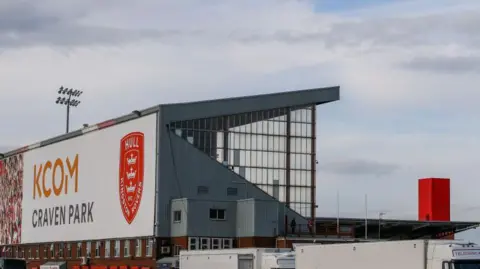 A general view of the exterior of Craven Park. There is a large metal stand in the foreground bearing the name of the stadium, along with a red tower and floodlight in the distance. The pitch is not visible.