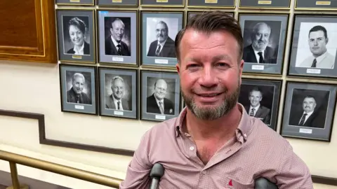 Sir Lee Pearson is stood wearing a red shirt with crutches to assist him. There are black and white photographs of former council leaders behind him.