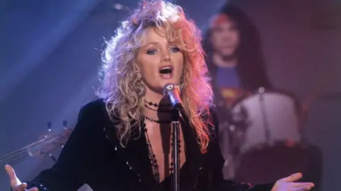 Bonnie Tyler singing into a microphone. She has big blonde hair and a black dress on. Behind is a drummer - he is slightly blurred, as Bonnie is the focus of the image.