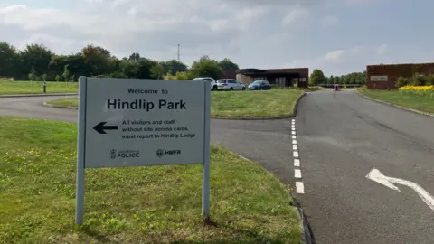 A white sign with two legs is positioned on a stretch of grass near a private road. The white sign reads: "Welcome to Hindlip Park." 