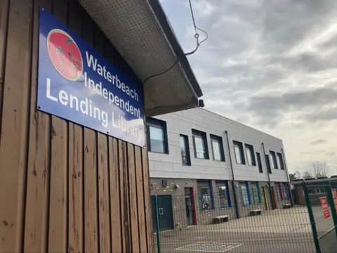 Jenny Kirk/BBC Side view of the library which shows a wooden wall with a sign saying Waterbeach Independent lending library. A modern school building is to the right of the library