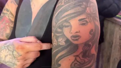 A heavily tattooed woman points to a tattoo of a woman's face on her upper arm.