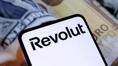 Reuters Фотография мобильного телефона с названием бренда Revolut и банкнотами на заднем плане за устройством.