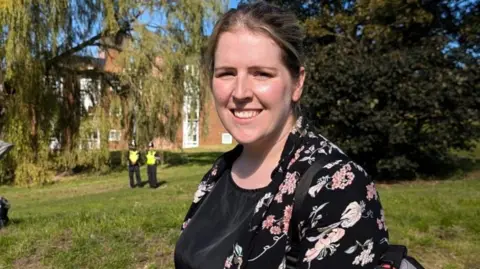 Rosalyn Ridley, who has light brown hair worn in a ponytail and wears a black patterned jacket, stands by some trees. Two police officers can be seen in the background.