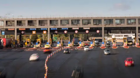 LCRCA A set of toll booths with cars passing through barriers underneath a grey concrete structure containing control rooms. 