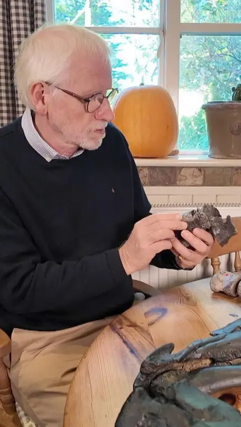 A white haired man wearing a blue jumper examines a dinosaur bone in his hands while sitting in his kitchen at the table