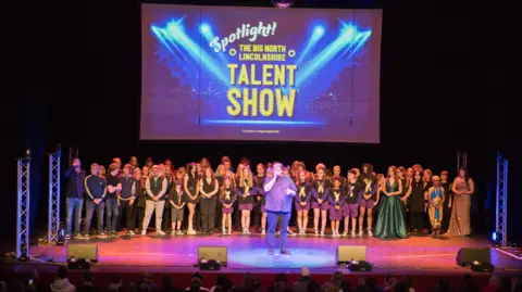 Scunthorpe Theatres A large group of people stand on stage, with a man in a purple shirt singing into a microphone at the front under a spotlight. Behind them, there is a screen that reads "Spotlight! The Big North Lincolnshire Talent Show", in bold, yellow font.