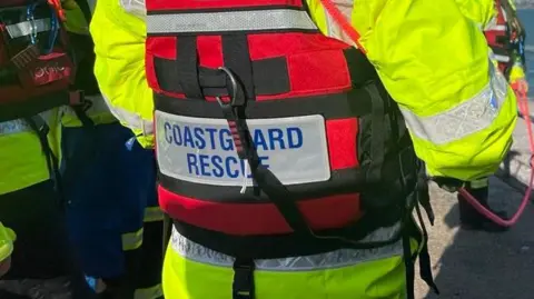 The back of a coastguard vest. The person is wearing a hi-vis jacket with a red lifejacket with the words 'Coastguard Rescue' on the back.