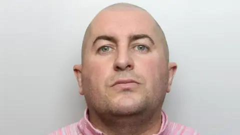 A police mughsot shows a man with a shaved head in a pink top against a grey background.