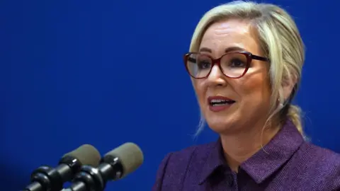 First Minister of Northern Ireland Michelle O'Neill speaks during a press conference, she is wearing a purple blazer and glasses. She has blonde hair.