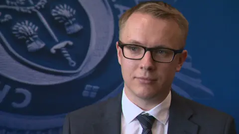  PC Keith Terrill, who is wearing a grey suit, white shirt and black striped tie,is staring off camera as he is being interviewed in this candid shot. 