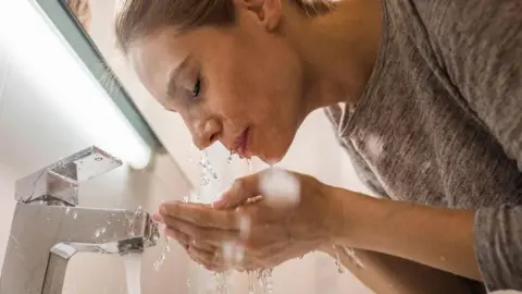 Woman with her head next to a running tap splashes water over her face with her hands. 