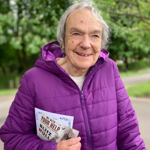 A woman with short grey hair is stood in front of trees and smiles at the camera. She's wearing a purple, hooded, zipped jacket. She has a pamphlet in her hand which reads "Blitz in Hull".