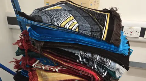 Razia Hadait Mats of various colours are piled up from top to bottom. Two electrical sockets are on the far right of the photo.