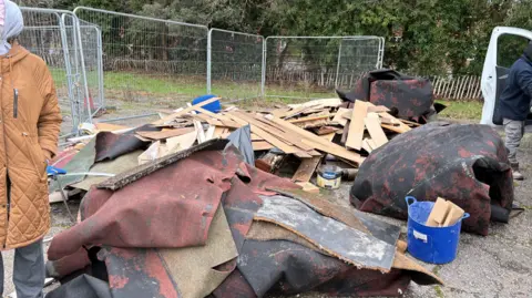Daniel Berkoh-Gyamfi Planks of wood and discarded rugs placed near a green space. The is a person partially in view to the right of the image. 