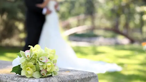Stock image shows a bunch of flowers on a stone in front of an anonymous bride and groom standing outside on a sunny day on grass in front of a little bridge feature in a garden.