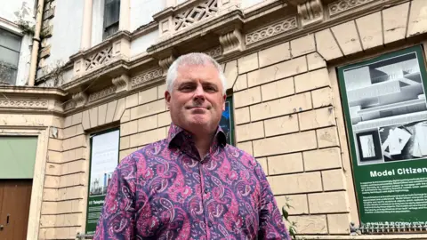 A man stands in front of the beige assembly buildings. He wears a pink and purple shirt. The man has short grey hair. There is a green notice board in place of a window over his left shoulder. 
