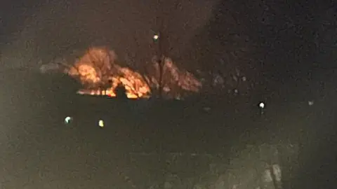 Kate Evans Flames from a fire on Rosenannon Downs head towards the night's sky. The foreground is dark but a set of buildings and trees can be made out.