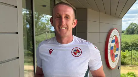 Reading FC Ed Jackson-Norris stands in a white t-shirt with the Reading FC badge. In the background a large Reading FC club crest is visible on the side of a building. 