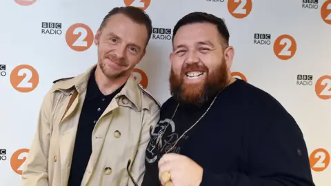 Simon Pegg (L) and Nick Frost (R) are smiling in a photo with BBC Radio 2 logos in the background. Simon is wearing aa beige trench coat and Nick is wearing a black jumper with a gold chain, gold ring and holding sunglasses. 