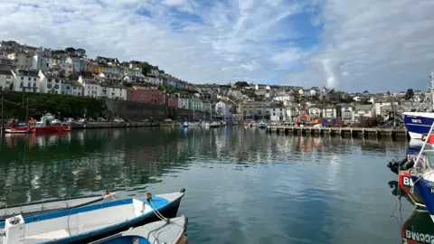 Brixham Harbour with calm water, small boats in the foreground, and a hillside of colourful houses under a partly cloudy sky.
