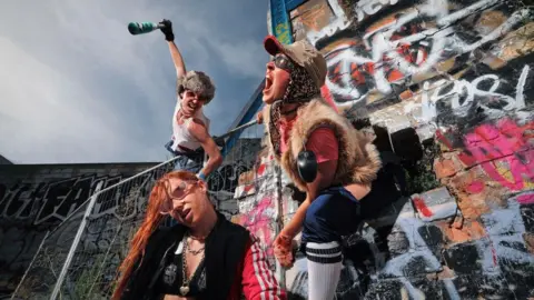 Matt Hurley Three people wearing extravagant outfits pose in front of a graffiti-covered wall under a hazy blue sky. One man is wearing sunglasses, a beige cap, furry gilet, pink top and white socks and is shouting. Another man, wearing sunglasses, a furry hat and white vest, appears to be diving towards a woman while holding a bottle. The woman has her head tilted to one side, as if listening. She has ginger hair and is wearing sunglasses, a black bra, black top and red tracksuit.