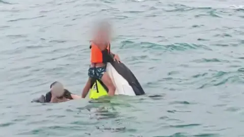 A boy wearing a lifejacket holds on to the top of a capsized jet ski as a man swims next to him in the sea