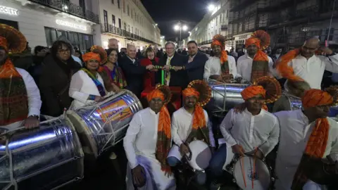 Dozens people gather to celebrate Diwali. A number of men are sat at the front wearing white collared shirts and orange head coverings.